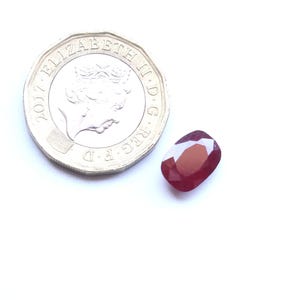 May include: A faceted, oval-shaped ruby gemstone with a deep red hue, next to a United Kingdom one-pound coin. The coin is silver and features a portrait of Queen Elizabeth II. The gemstone is approximately 1.3 cm in length.