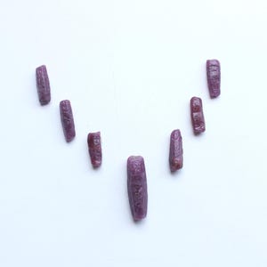 Pu&ograve; includere: Una collezione di gemme di rubino grezze, non tagliate, ciascuna con una tonalit&agrave; rosso-viola intenso. Le pietre sono disposte su una superficie bianca, mostrando le loro forme e texture naturali e irregolari. Adatte per la creazione di gioielli.