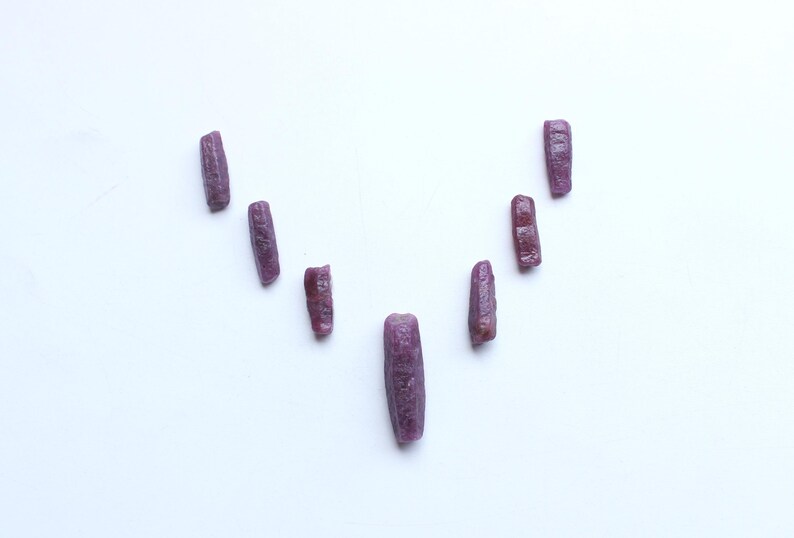 Pu&ograve; includere: Una collezione di gemme di rubino grezze, non tagliate, ciascuna con una tonalit&agrave; rosso-viola intenso. Le pietre sono disposte su una superficie bianca, mostrando le loro forme e texture naturali e irregolari. Adatte per la creazione di gioielli.