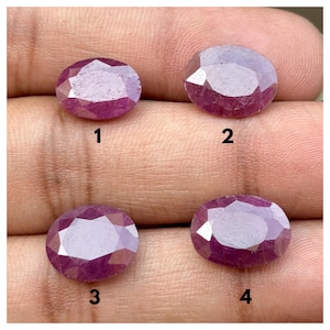 Puede incluir: Cuatro piedras preciosas ovaladas y facetadas de color rojo rosáceo intenso dispuestas en cuadrícula. Cada piedra está etiquetada con un número negro del uno al cuatro. Las piedras muestran una superficie pulida y reflectante con inclusiones.
