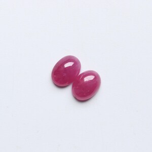 May include: Two oval-shaped, polished gemstones in a vibrant, deep pink hue. The gems are smooth and glossy, reflecting light. They are set against a plain white background, highlighting their colour and shape. These could be used for jewellery making.