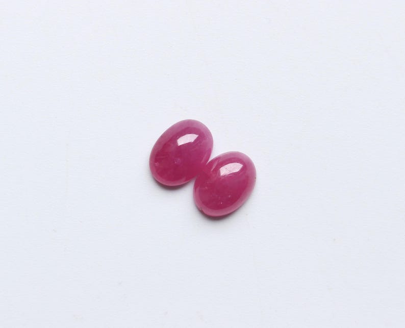May include: Two oval-shaped, polished gemstones in a vibrant, deep pink hue. The gems are smooth and glossy, reflecting light. They are set against a plain white background, highlighting their colour and shape. These could be used for jewellery making.