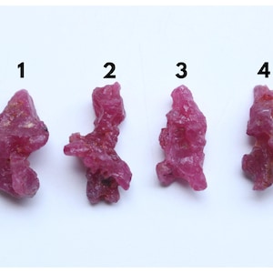 May include: Four rough, uncut ruby gemstones, numbered 1 through 4, are displayed against a white background. The rubies are a deep, vibrant red color, with irregular shapes and textured surfaces. They appear to be natural, unpolished specimens.