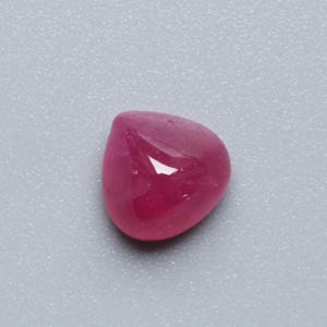 May include: A polished, cabochon-cut ruby gemstone. The gem is a deep, saturated red colour with a smooth, rounded shape. The surface reflects light, creating highlights. The ruby is set against a light grey background.