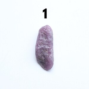 May include: A rough, elongated, purple gemstone with a textured surface. The stone is set against a white background, with the number "1" in black at the top. The gemstone's colour varies from light to dark purple.