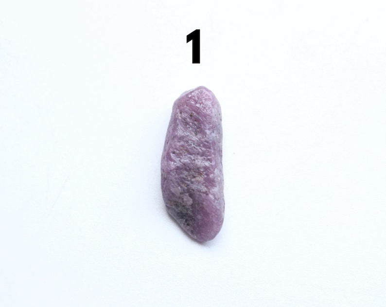 May include: A rough, elongated, purple gemstone with a textured surface. The stone is set against a white background, with the number "1" in black at the top. The gemstone's colour varies from light to dark purple.