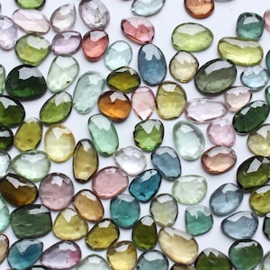 Op de afbeelding: Een verzameling gefacetteerde edelstenen in verschillende kleuren, waaronder groen, roze, geel en blauw. De edelstenen zijn ovaal en druppelvormig, met een gepolijst, reflecterend oppervlak. De edelstenen zijn verspreid over een witte ondergrond.