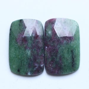 Puede incluir: Dos piedras preciosas rectangulares con tapas facetadas. Las piedras tienen un color base verde con inclusiones rojas y moradas. Las piedras preciosas están sobre un fondo blanco.