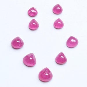 May include: Nine teardrop-shaped, polished ruby gemstones. The rubies are a vibrant pinkish-red colour and are arranged in a circular pattern against a white background. Each stone has a smooth, rounded surface.