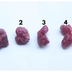 May include: Four raw, unpolished ruby gemstones arranged in a horizontal row. Each stone displays a deep pinkish-red hue with irregular, jagged textures and natural crystalline facets. The stones are labeled with the black numbers 1, 2, 3, and 4.