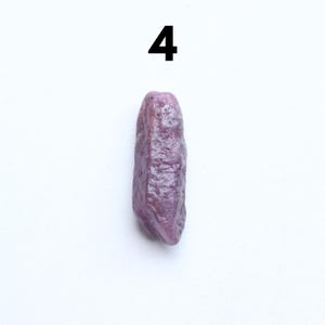 May include: A rough, elongated, purple gemstone with a textured surface. The number "4" is printed in black above the gemstone against a white background. The gemstone is a natural, unpolished mineral.