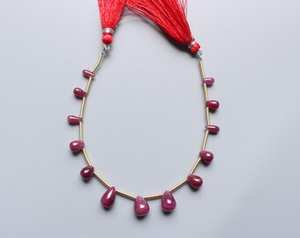 May include: A necklace of ruby gemstone beads on a gold-coloured wire. The beads are a deep red colour and teardrop-shaped. Red tassels are attached to the top of the strand. The background is a light grey colour.