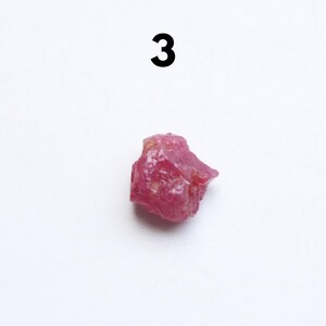 Puede incluir: Una gema de rub&iacute; en bruto, sin cortar, con un vibrante tono rojo-rosado. La piedra natural tiene una forma irregular y una superficie texturizada. El n&uacute;mero "3" est&aacute; impreso en negro.