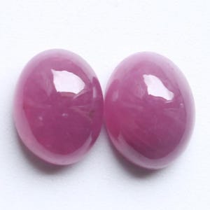 Op de afbeelding: Twee ovale, gepolijste edelstenen met een dieproze tint. De stenen hebben een glad, glanzend oppervlak dat licht reflecteert. De edelstenen zijn geplaatst tegen een witte achtergrond.