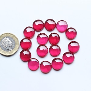 May include: A collection of sixteen round, polished, ruby-red gemstones arranged in a circle. A gold coin is positioned near the gemstones for size comparison. The gemstones have a glossy, reflective surface.