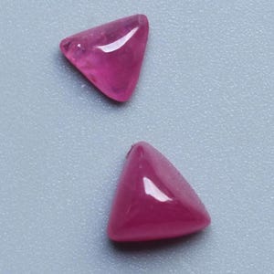 May include: Two triangular, polished gemstones in a vibrant magenta hue. The gems have a smooth, rounded surface and a glossy finish, set against a light grey background. These gemstones are likely for jewellery making or collecting.