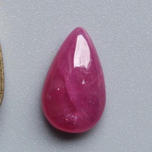 Op de afbeelding: Een gepolijste, druppelvormige robijn edelsteen. De edelsteen heeft een diepe, verzadigde roze-rode kleur met een glad, reflecterend oppervlak. De steen ligt tegen een neutrale grijze achtergrond.