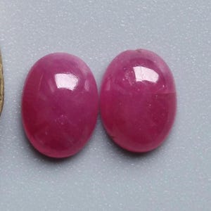 May include: Two oval-shaped, polished gemstones in a vibrant magenta hue. The stones have a smooth, reflective surface, catching the light. The gemstones are set against a light grey background.