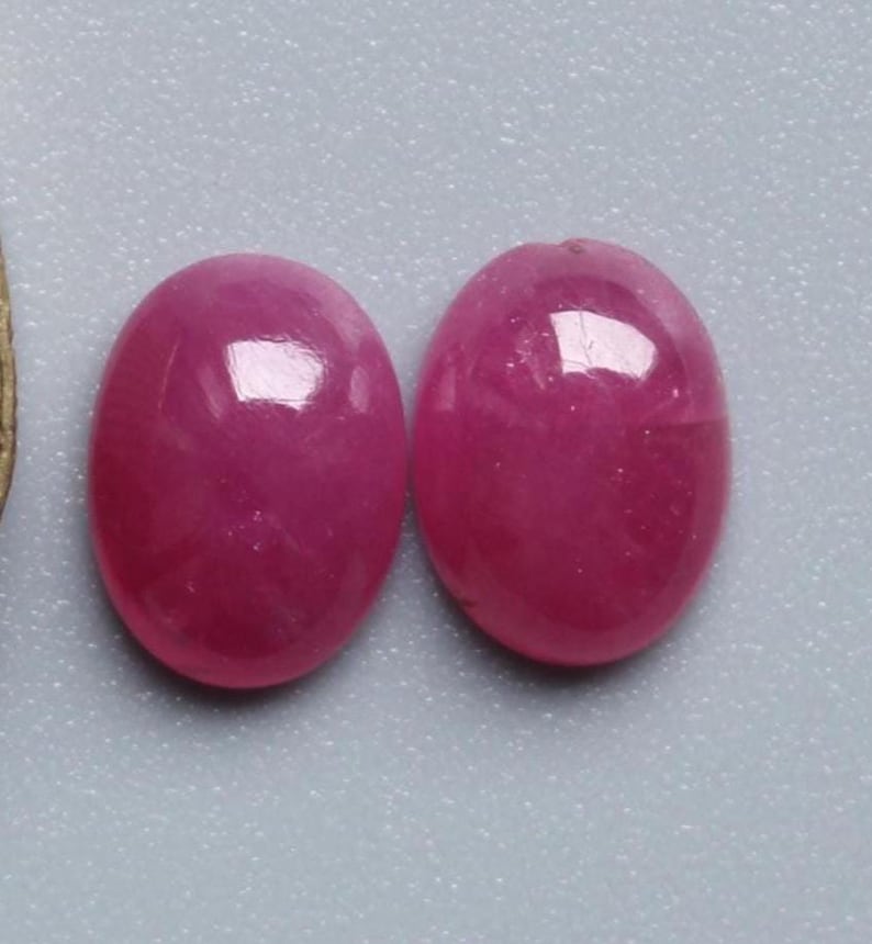May include: Two oval-shaped, polished gemstones in a vibrant magenta hue. The stones have a smooth, reflective surface, catching the light. The gemstones are set against a light grey background.