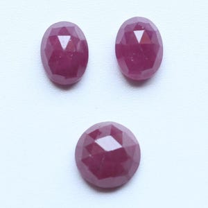 Puede incluir: Tres piedras preciosas facetadas de color magenta rosado intenso. Dos de las piedras tienen forma ovalada, mientras que la tercera es redonda. Cada piedra presenta una superficie geométrica de talla rosa con múltiples facetas.