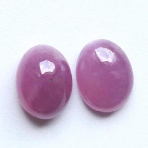 Peut inclure: Deux pierres précieuses de forme ovale, taillées en cabochon, avec une surface lisse et polie d'une teinte rose-violet éclatante. Chaque pierre présente une finition brillante et réfléchissante qui capte la lumière, soulignant la couleur uniforme.