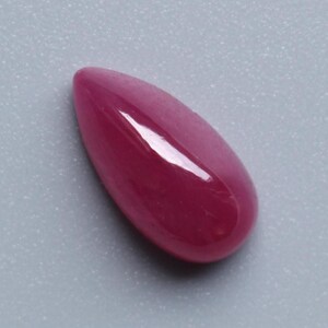 Op de afbeelding: Een gepolijste, druppelvormige edelsteen in een diepe, rijke magenta tint. Het gladde oppervlak reflecteert licht en creëert highlights. De edelsteen ligt tegen een lichtgrijze achtergrond. Geschikt voor het maken van sieraden.