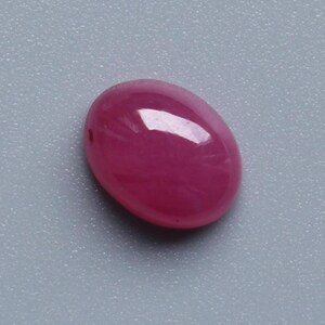 May include: A polished, oval-shaped ruby gemstone. The gem is a deep, saturated pinkish-red colour with a glossy surface. The stone is set against a light grey background.