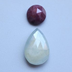 May include: Two gemstones: a round, faceted, dark red ruby and a pear-shaped, faceted, pale blue aquamarine. The aquamarine has a soft, milky appearance. Both stones are set against a light blue-gray background.