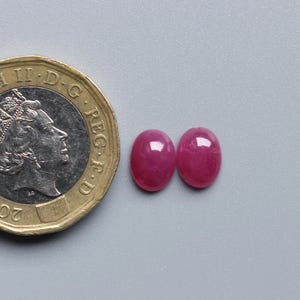 Op de afbeelding: Twee ovale, helderrode edelstenen naast een Britse munt van &eacute;&eacute;n pond. De edelstenen hebben een gepolijst oppervlak. De munt is goud en zilver van kleur. De edelstenen zijn ongeveer 1 cm lang.
