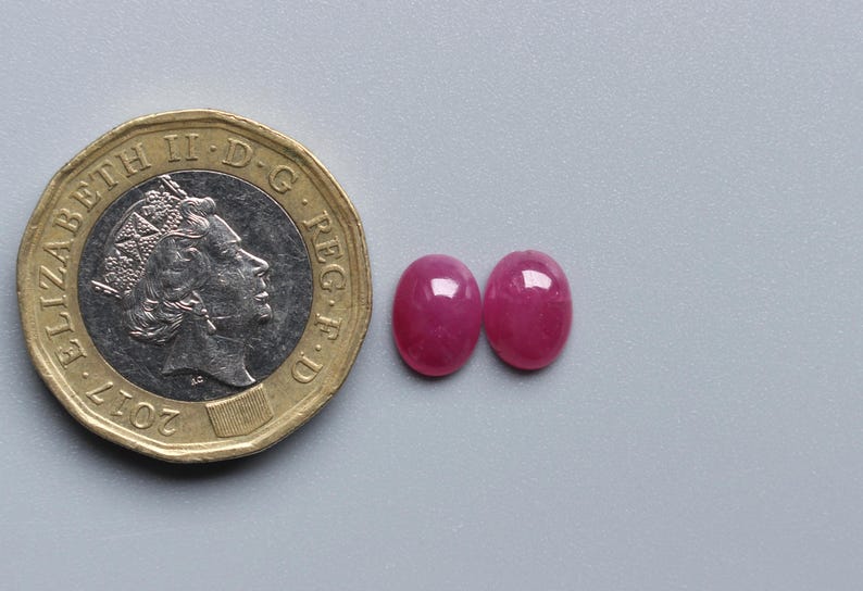 Op de afbeelding: Twee ovale, helderrode edelstenen naast een Britse munt van &eacute;&eacute;n pond. De edelstenen hebben een gepolijst oppervlak. De munt is goud en zilver van kleur. De edelstenen zijn ongeveer 1 cm lang.