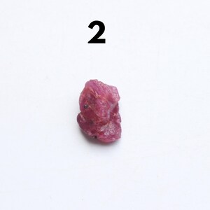 Puede incluir: Una gema de rub&iacute; en bruto, sin cortar, de color rojo intenso. La piedra natural tiene una forma irregular y una superficie ligeramente texturizada. El n&uacute;mero "2" est&aacute; impreso en negro encima de la gema.