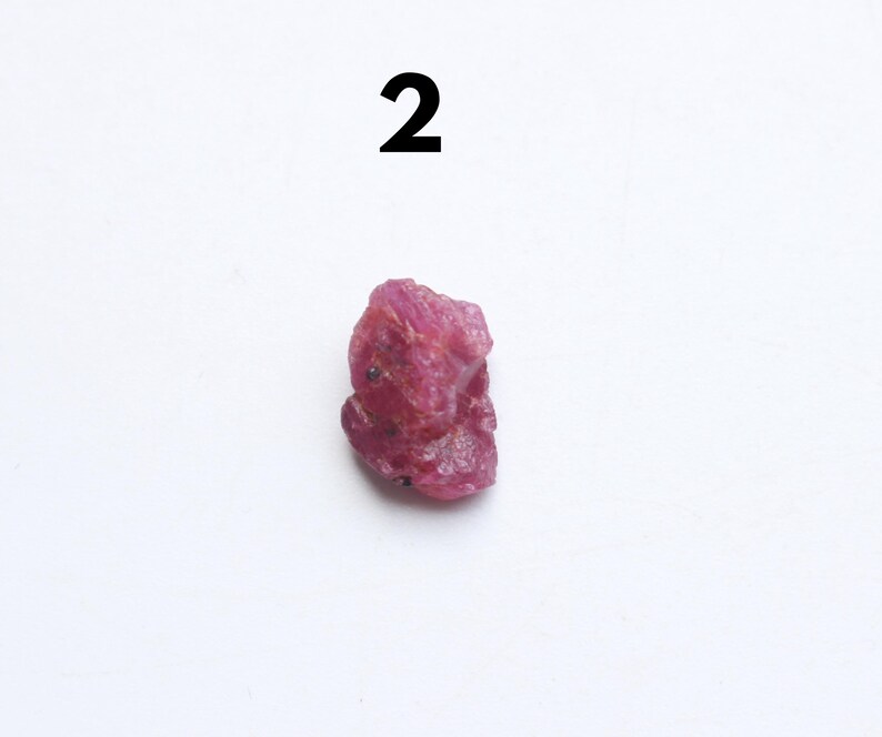 Puede incluir: Una gema de rub&iacute; en bruto, sin cortar, de color rojo intenso. La piedra natural tiene una forma irregular y una superficie ligeramente texturizada. El n&uacute;mero "2" est&aacute; impreso en negro encima de la gema.