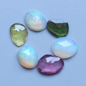 Op de afbeelding: Een verzameling gefacetteerde edelstenen in verschillende vormen en kleuren. De edelstenen omvatten opalen, een groene steen en een paarse steen. De stenen zijn gerangschikt op een lichtblauwe ondergrond, waarbij hun unieke slijpsels en kleuren worden getoond.