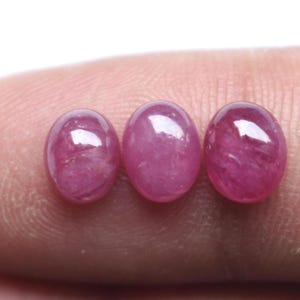 May include: Three oval-shaped, cabochon-cut pink rubies with a smooth, polished surface. Each gemstone displays a translucent quality with internal inclusions and a reflective luster, arranged in a horizontal row against a neutral background.