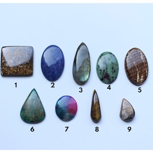 May include: Nine different gemstone cabochons in various shapes and colors, including brown, blue, green, and gray. The cabochons are numbered 1 through 9.