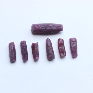 Pu&ograve; includere: Una collezione di gemme di rubino grezze e non tagliate. Le gemme sono di un colore rosso-violaceo intenso e hanno una superficie ruvida e strutturata. Una pietra pi&ugrave; grande &egrave; sopra sei pietre pi&ugrave; piccole.