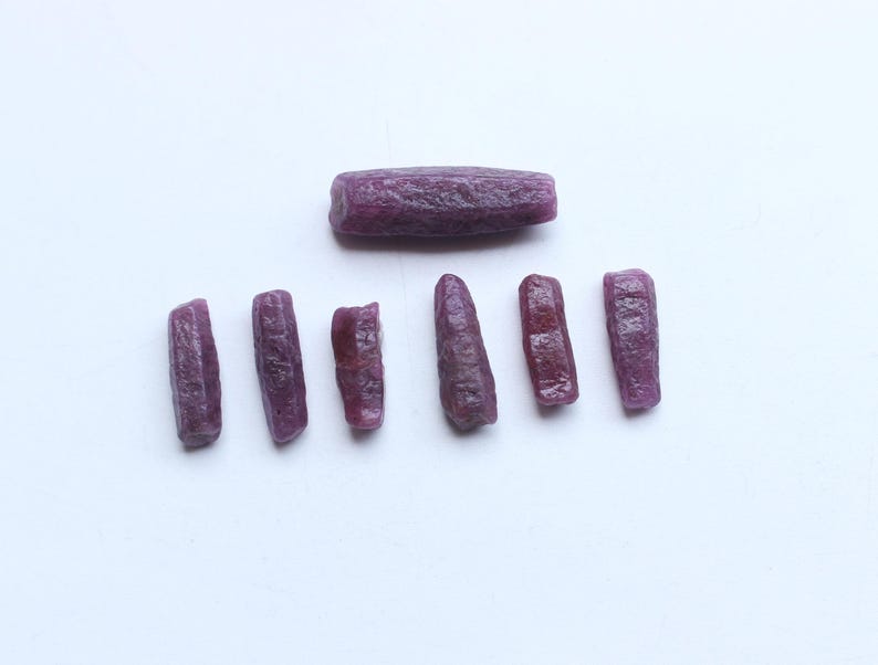 Pu&ograve; includere: Una collezione di gemme di rubino grezze e non tagliate. Le gemme sono di un colore rosso-violaceo intenso e hanno una superficie ruvida e strutturata. Una pietra pi&ugrave; grande &egrave; sopra sei pietre pi&ugrave; piccole.