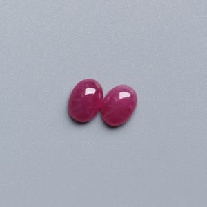 May include: Two oval-shaped, polished gemstones in a vibrant, deep pink hue. The gems have a smooth, reflective surface, catching the light. The background is a solid, muted grey, providing a neutral backdrop for the gemstones. These are likely loose gemstones for jewellery making.