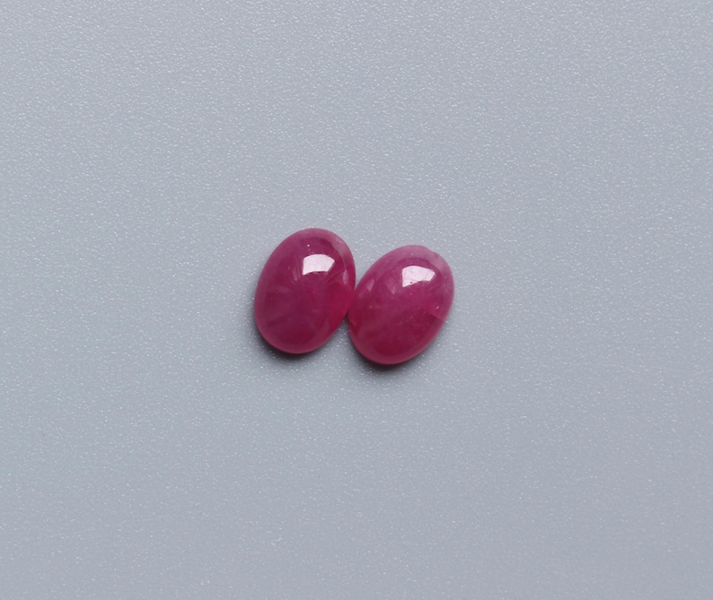 May include: Two oval-shaped, polished gemstones in a vibrant, deep pink hue. The gems have a smooth, reflective surface, catching the light. The background is a solid, muted grey, providing a neutral backdrop for the gemstones. These are likely loose gemstones for jewellery making.