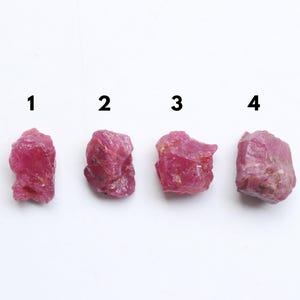 Puede incluir: Cuatro piedras preciosas de rubí en bruto y sin cortar se muestran sobre un fondo blanco. Las piedras preciosas están numeradas del 1 al 4. Cada rubí tiene un color rojo rosado intenso y una textura rugosa e irregular.