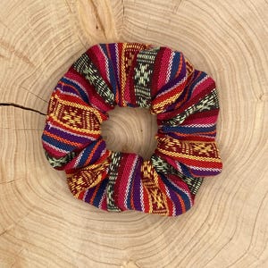 May include: A colorful scrunchie with a woven pattern in red, purple, yellow, and blue. The hair accessory is made of fabric and is circular in shape. The scrunchie is displayed on a wooden surface.