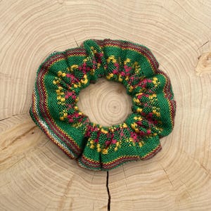 Può includere: Un elastico per capelli verde con un motivo intrecciato di disegni floreali gialli e rosa. L'elastico per capelli ha un bordo a righe rosse e bianche. L'accessorio per capelli è circolare e poggia su una superficie di legno.