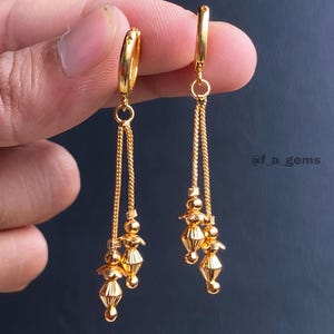 May include: A pair of gold-toned hoop earrings featuring two delicate dangling chains. Each chain ends with a small, textured, bead-like charm. The text @f_a_gems is visible on the right side. These earrings have a polished, reflective finish.
