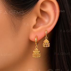 Könnte beinhalten: Ein Paar goldene Creolen mit baumelnden, glockenförmigen Jhumka-Anhängern, die mit filigranen Metallarbeiten und kleinen Perlen an den unteren Rändern verziert sind. Die Ohrringe werden am Ohrläppchen getragen und haben ein poliertes Gelbgold-Finish.