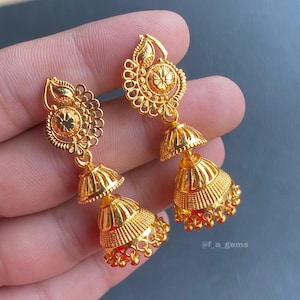 May include: A pair of gold-colored earrings with a floral design. The earrings have a detailed, ornate pattern with a bell-shaped bottom and a decorative top. The earrings are held in a hand, showcasing their intricate craftsmanship.