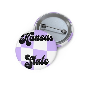 Puede incluir: Una chapa redonda con un patrón de cuadros en color violeta claro y blanco. El texto Kansas State está impreso en una fuente negra, negrita y estilizada en el centro. La parte posterior muestra un mecanismo de alfiler de metal plateado.