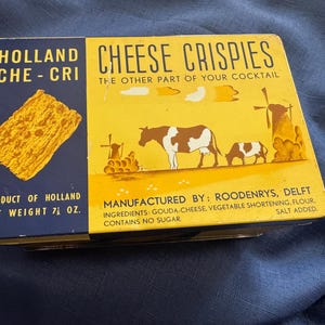May include: Vintage Holland Cheese Crispies tin. The rectangular tin features a yellow and blue design with the text "Cheese Crispies" and "Product of Holland." The tin has a net weight of 7.5 oz. and includes illustrations of cows and windmills.