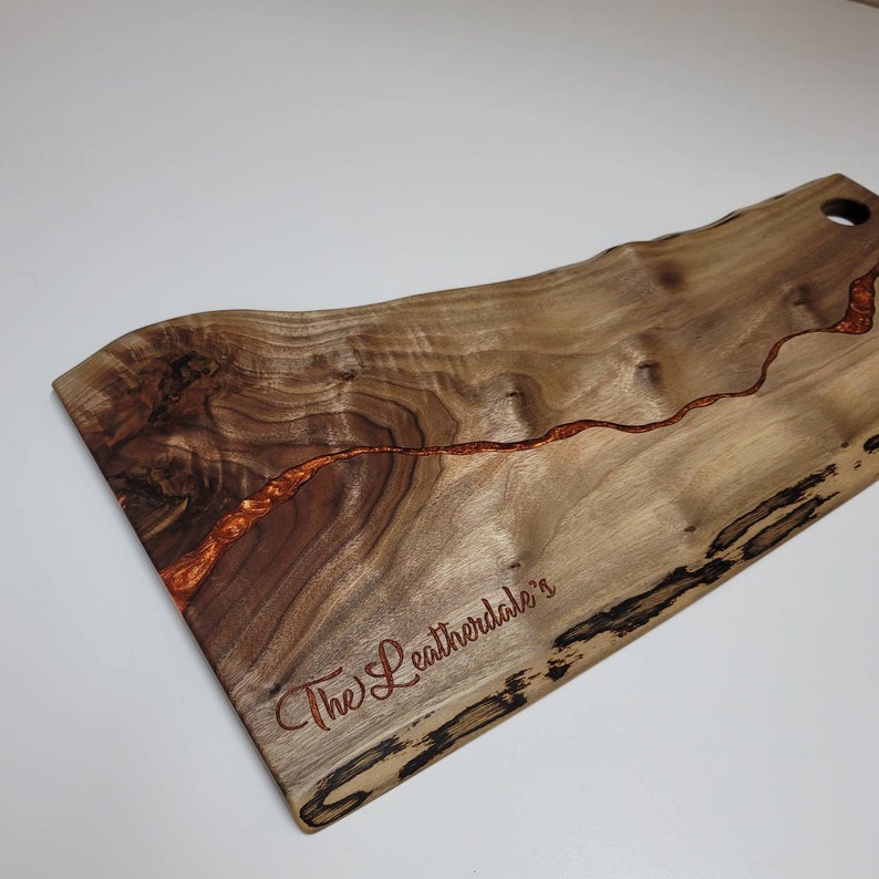 May include: A wooden serving board with a natural wood grain finish and a copper-colored epoxy resin inlay. The board has a live edge and is engraved with the text "The Leatherdale's".