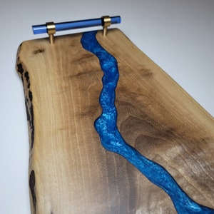 May include: A wooden serving board with a blue resin river running through the center. The board has a live edge and a brass handle with a blue glass insert.