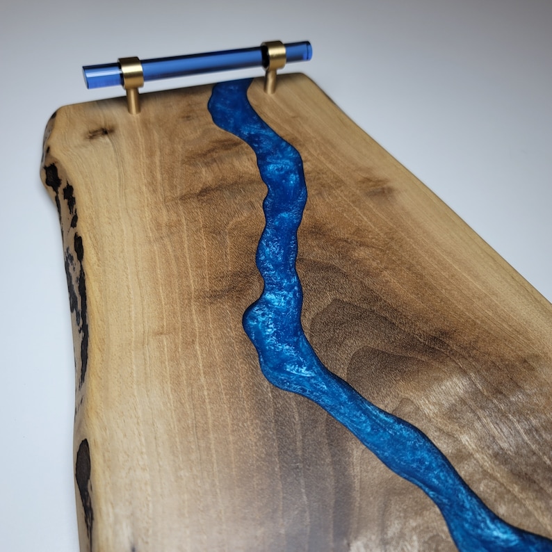 May include: A wooden serving board with a blue resin river running through the center. The board has a live edge and a brass handle with a blue glass insert.